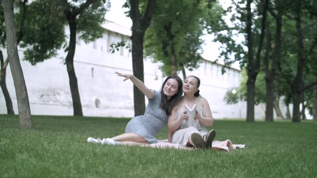 Happy smiling mother and daughter reading book in sunny park outdoors. Positive woman and girl enjoying leisure in sunshine. Hobby and lifestyle concept. High quality FullHD footage
