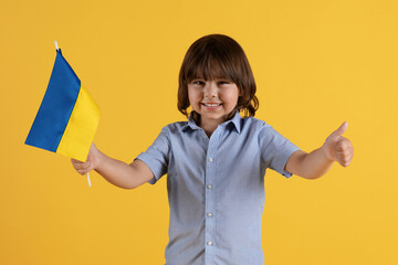 Happy cute little boy holding Ukrainian flag and showing thumb up, smiling widely to camera, orange background