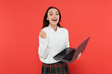 Excited woman in warm jumper showing yes gesture and holding laptop isolated on red