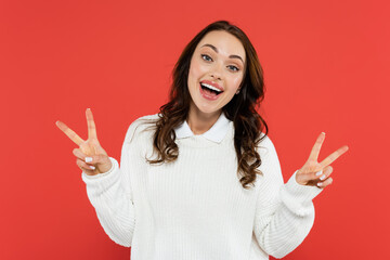 Positive brunette woman in white jumper showing peace sign isolated on red