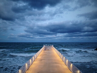 illuminated pier with dramatic sky over stormy dark sea at evening