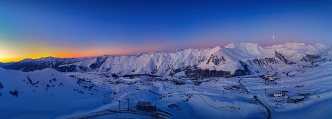 Wide aerial panorama of snowy mountain ridge on winter sunrise. Stunning mountains range covered with snow powder on ski resort at sunset. Moon above caucasus peaks skyline in the dusk at night. - obrazy, fototapety, plakaty