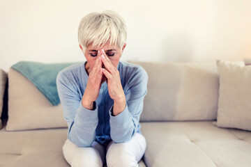 Sad depressed woman at home sitting on the couch, looking down and touching her forehead, loneliness and pain concept. Depressed and sad grey hair mature woman sitting on bed at home during the day.
