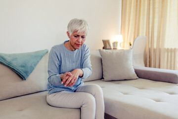 Frustrated stressed middle aged female retiree having painful feelings in bones, arthritis osteoporosis concept. Shot of an woman sitting alone on sofa at home and suffering from arthritis in her hand