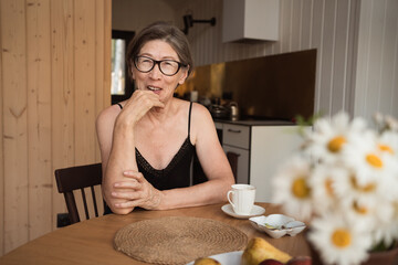 Happy senior 60s woman wearing glasses sitting at the kitchen table