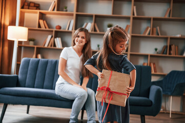 Fototapeta premium Girl holding gift box behind her. Happy holidays. Young mother with her daughter is together at apartment