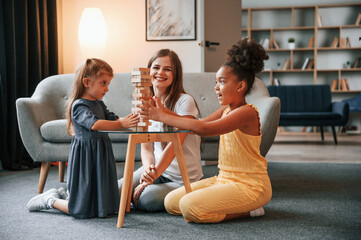 On the floor together. A young woman with two girls is playing a wooden tower game indoors