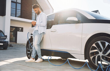 Standing with smartphone and waiting for charge to finish. Young stylish man is with electric car at daytime