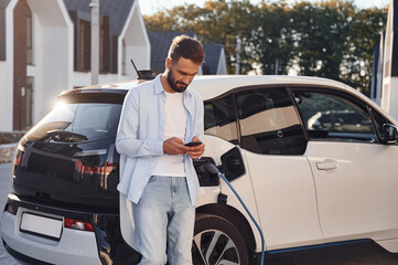 Waiting for the charge completes. With smartphone in hands. Young stylish man is with electric car...