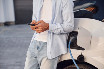 Standing with smartphone while automobile is charging. Close up view of man with his electric car