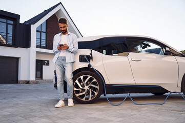 Beautiful young stylish man is with electric car at daytime charging the vehicle and holding smartphone