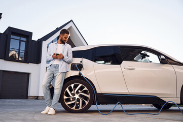 Standing and looking at vehicle that is being charging. Young stylish man is with electric car at...