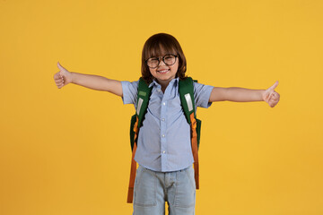 Adorable little boy preschooler in eyeglasses with backpack gesturing thumbs up with both hands,...