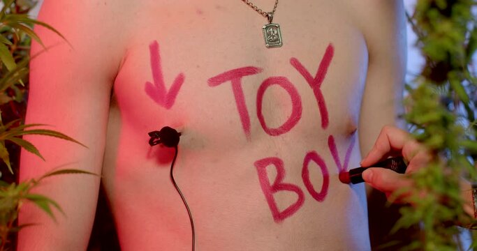 The inscription on the body of the boy toy. The hand writes letters with red lipstick. A microphone is attached to the body. Red, blue tinted colors.