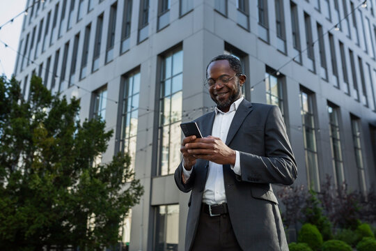 Mature african american boss using phone, businessman outside office building in business suit and glasses, senior man typing message and browsing online pages smiling.