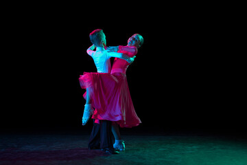 Waltz. Full-length portrait of beautiful little boy and girl dancing ballroom dance isolated over dark background in neon light. Concept of art, beauty, grace, action, emotions. - obrazy, fototapety, plakaty