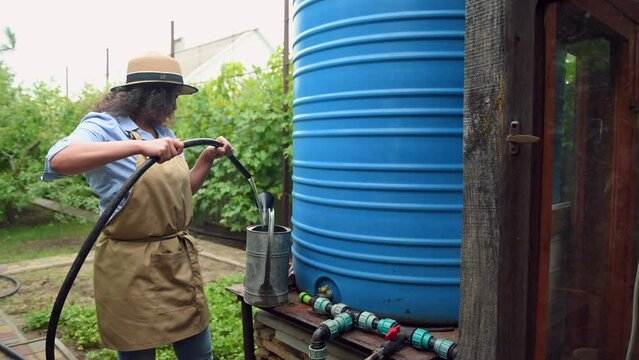 Multi-ethnic woman farmer, agriculturist gardener in a straw hat, collecting water from barrel to watering can for watering the garden and orchard. Gardening Farming Agricultural hobby and business