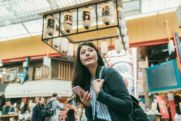 cheerful asian girl using cellphone for online information and looking around enjoying active vibe in kuromon ichiba market in Osaka japan. translation:â kuromon ichiba marketâ