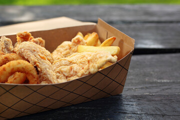Deep fried onion rings and breaded chicken strips, served with fries, in a takeaway box. Fast food, food truck meal.