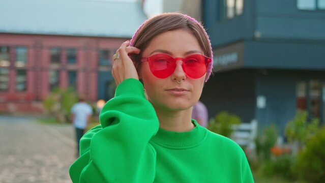 Close-up portrait of friendly-looking young woman in green sweatshirt smiling to camera in background of blurred urban view. Shot of cute girl wearing fashionable outfit. Red glasses. City, outdoor