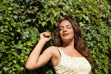Beautiful young woman with long brown hair and cinnamon skin leaning against a background of green ivy, eyes closed sunbathing. Concept beauty, fashion, make-up, relax, peace, plants.