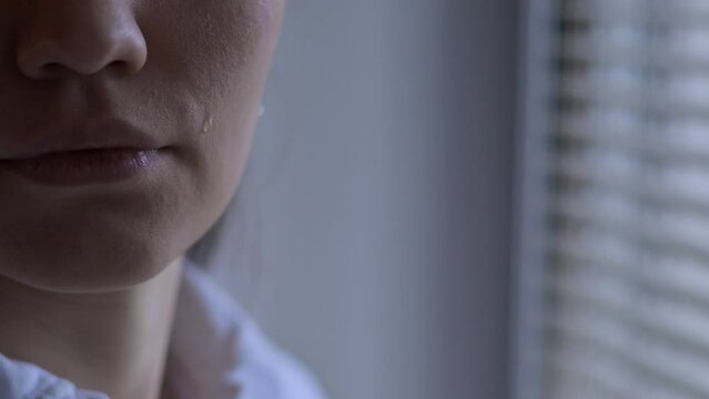 Close-up of a tear that rolls down the cheek of a woman who is standing near the window. The emotion of experiencing what is happening in the world - wars, disasters, epidemics and catastrophes
