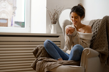 Smiling lady in smart trendy wear is sitting on armchair with a cup of tea. Smiling, sitting in relaxing atmosphere indoors at home, nice design.
