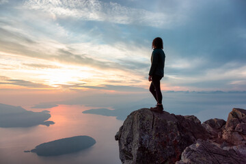 Adventurous Woman Hiker on top of Canadian Mountain Landscape on the West Coast of Pacific Ocean. Dramatic Sunset and Hazy Smoky Sky. St. Mark's Summit near Vancouver, British Columbia, Canada.