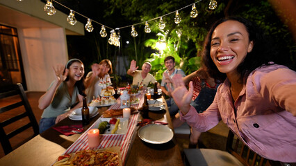 Authentic shot of happy carefree smiling woman making selfie or video call to friends or relatives while at garden party celebration with big family