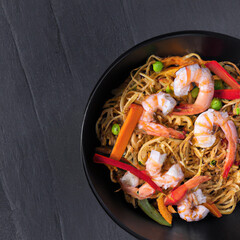 Stir fry noodles with vegetables and shrimps in black bowl. Slate background. Top view
