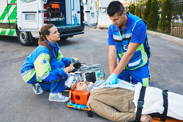EMS, first aid. Two paramedics performing closed-chest cardiac massage for victim man who lies unconscious on medical stretcher near ambulance outdoor © Peakstock