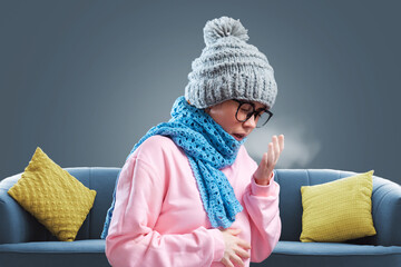 Young woman wearing glasses, in hat and knitted scarf is warming herself and cough. Gray background...