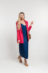 Fashion happy pretty blonde woman with smile in stylish outfit with blue long elegance dress, bright pink jacket and fashionable bag with shoes walks on a white background in studio