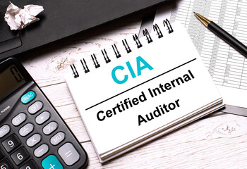 On a light-colored office table is a computer, a black calculator, a pen, and a notebook with the text CIA Certified Internal Auditor - obrazy, fototapety, plakaty
