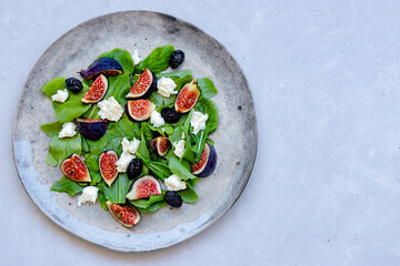 Fresh healthy summer salad with figs, white cheese, arugula and black olives on gray ceramic plate on gray background. Top down view. Close-up. Copy space.