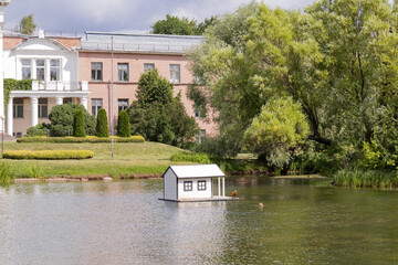 Pond, floating duck's house with ducks. Trees, bushes, pinkish building, landscape. Botanical...