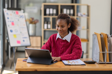 Smiling young African american female Finance Entrepreneur sitting in workplace office reading paperwork and working on a laptop, investment financial document report finance analysis concept.