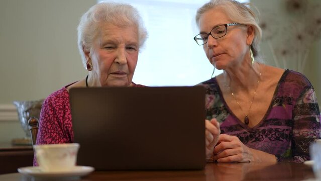 Elderly senior woman looking at computer with mature daughter next to her, both enjoying time together sharing and learning.