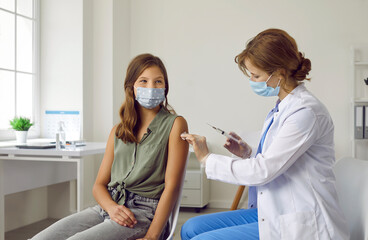 Vaccination procedure. Smiling brave teenage girl wearing mask receives dose of coronavirus vaccine at vaccination center. Female doctor holds syringe and disinfects injection site on child's hand.