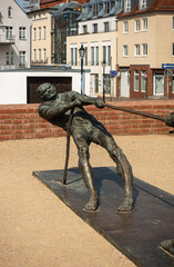 Naklejka premium Germany, Wismar - July 13, 2022: St. Marienkirche. Detail of 1 man at Tauziehen or tug-of-war statue in front. Multicolor house facades around square in back. Yellow gravel bottom