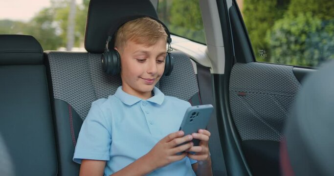 Good-looking satisfied teen boy in headphones sits in car's backseat and looking for modern music using his smartphone