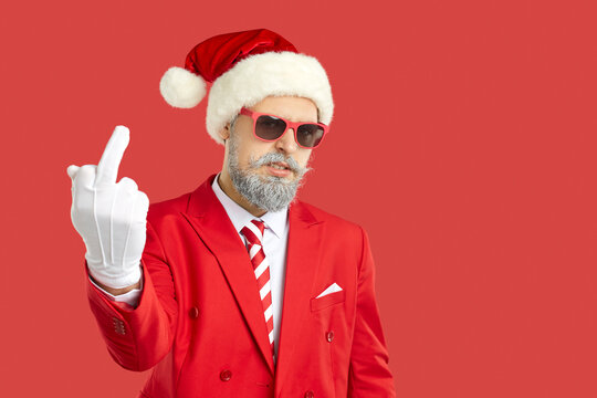 Bad rude Santa wishes you Merry Christmas season. Bearded man in red suit, tie, hat, cool sunglasses and white gloves, standing on red studio background, shows middle finger with angry face expression