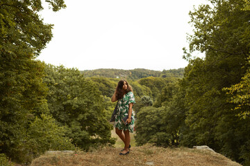 Fototapeta premium A young brunette woman with a green flower dress and a black book in her hand standing on the top of a hill on rocks in the forest in summer.