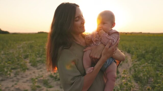 Cute mother lulling lovely child, kissing baby on field. Portrait of smiling young Caucasian woman holding toddler in hands in background of beautiful sunset. Summertime, evening outdoor