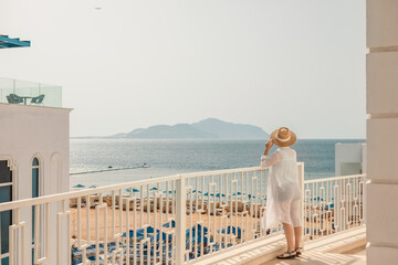 view from behind of a woman in a hat and a white shirt looking from the hotel balcony at the sea, the concept of a vacation at sea and travel