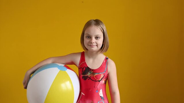 A girl in a red swimsuit is standing with a beach inflatable ball looking at the camera and laughing on a yellow background