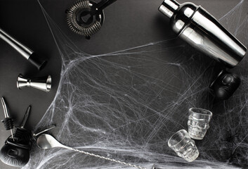 Various bar accessories such as: a shaker, muddler, strainer, as well as a bar spoon and a measuring stack lie against the backdrop of a spider web on a black matte table. Adult halloween celebration