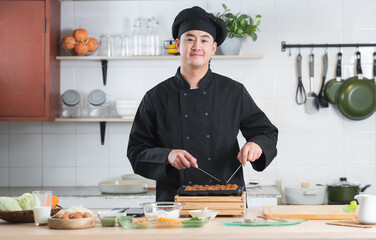 Portrait of handsome young Asian man chef wear black uniform smiling and standing, making japanese food called takoyaki with ingredients on table at kitchen restaurant
