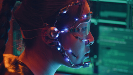 Fototapeta premium Side view of an Asian girl with futuristic headset and microphone programming using multiple computer screen. Cyperpunk style. Sci-fi background.