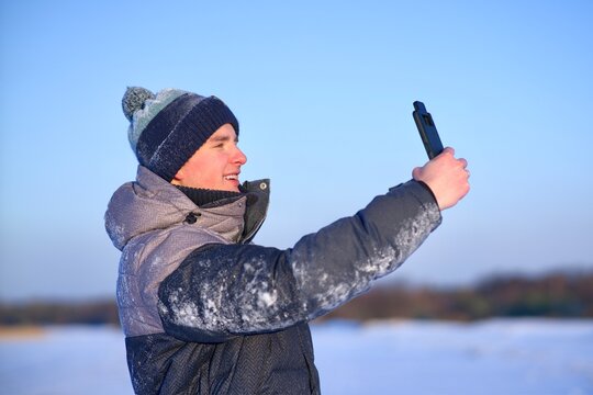 Happy handsome guy, young man blogger is taking selfie, picture of himself for social media on his cell mobile phone, looking at frontal camera of smartphone outdoors at winter cold frosty snowy day